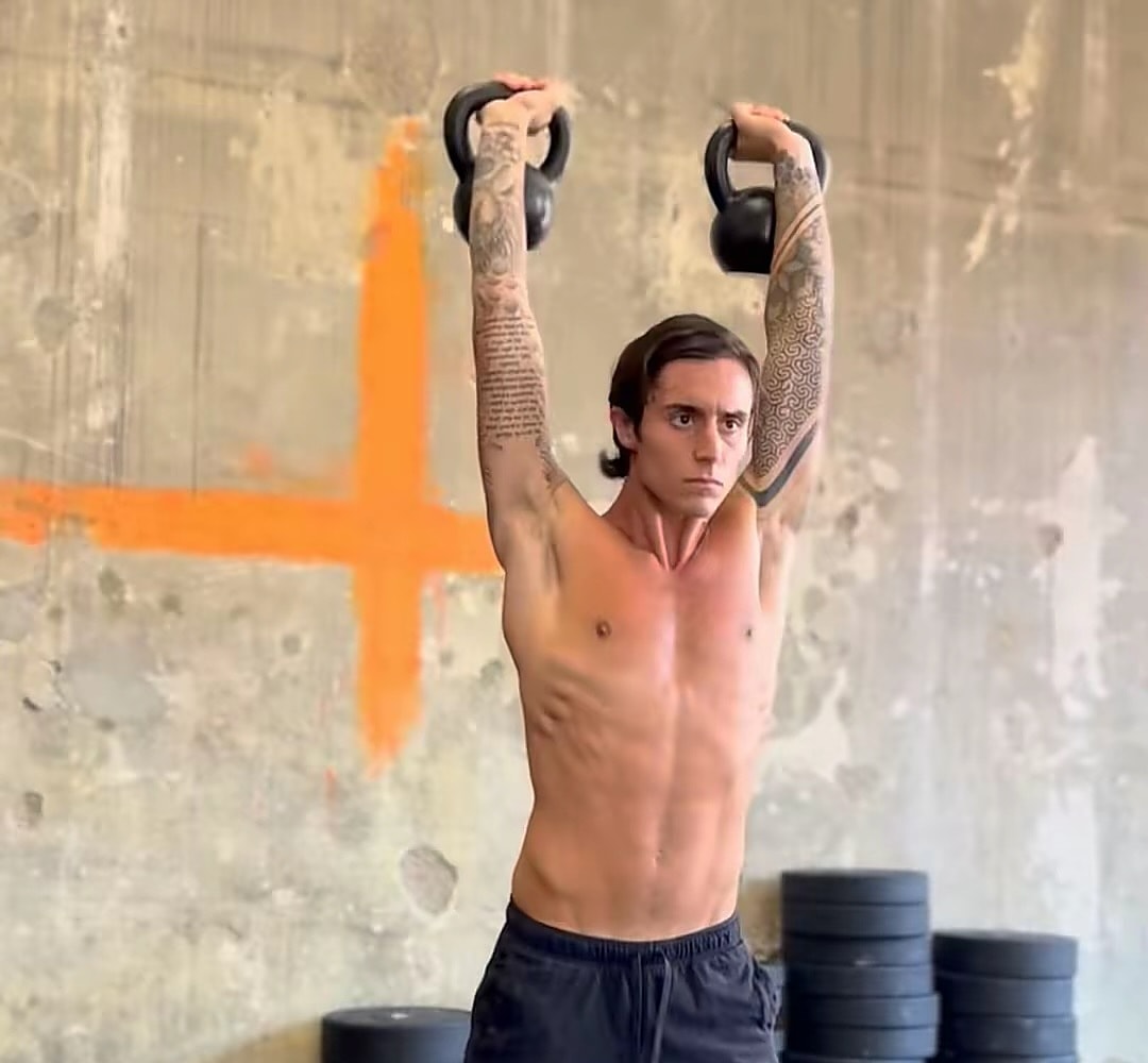Giuseppe in palestra con due kettlebell sopra la testa, davanti a un muro con una croce arancione dipinta
